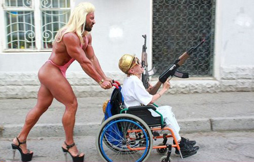 un jeune dguis en papy arm de mitraillettes dans un fauteuil roulant et pouss par un mec body-build, roi de la gonflette en dshabill rouge, talons haut compenss et avec une perruque blonde